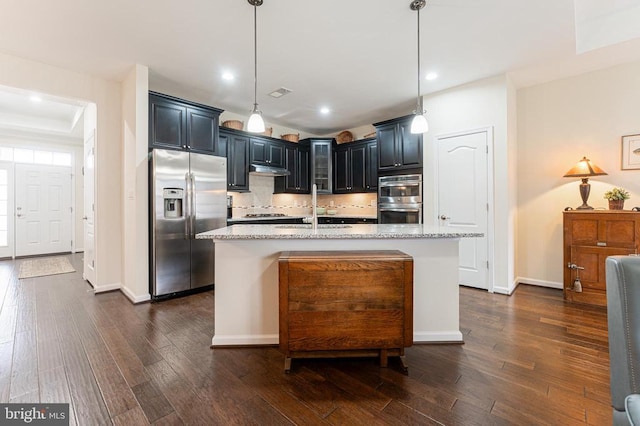 kitchen featuring light stone countertops, decorative light fixtures, stainless steel appliances, and an island with sink