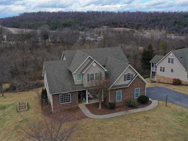 traditional-style house with driveway, a shingled roof, stone siding, a front lawn, and a wooded view