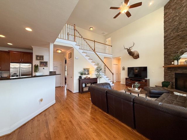 living area featuring light wood-style flooring, stairway, a high ceiling, a ceiling fan, and a stone fireplace