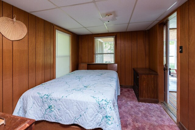 carpeted bedroom with a drop ceiling, multiple windows, and wood walls