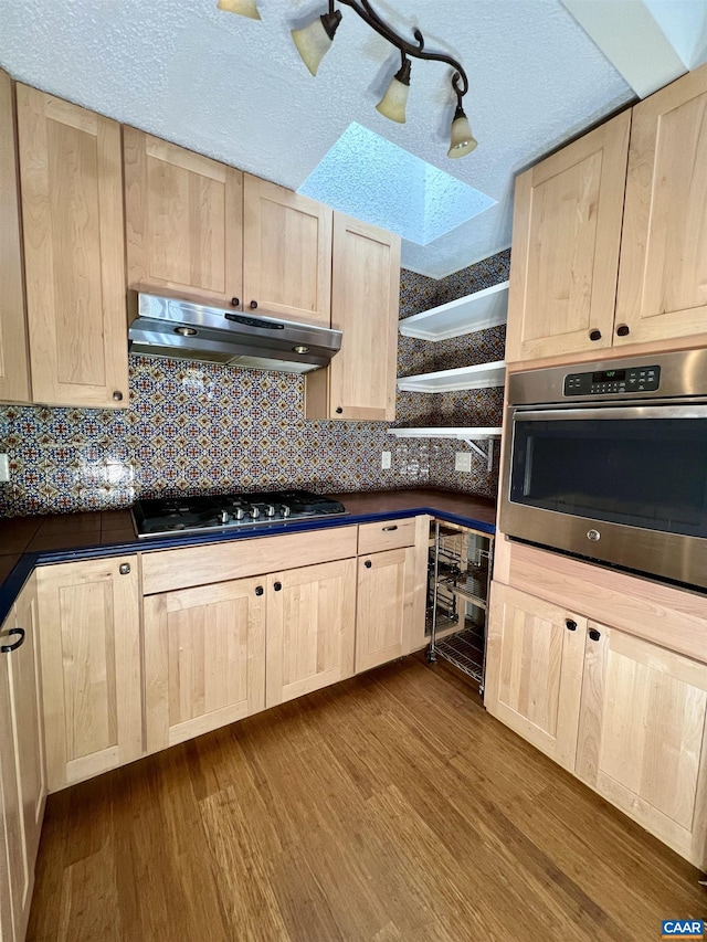 kitchen featuring stainless steel appliances, dark countertops, light brown cabinetry, beverage cooler, and under cabinet range hood