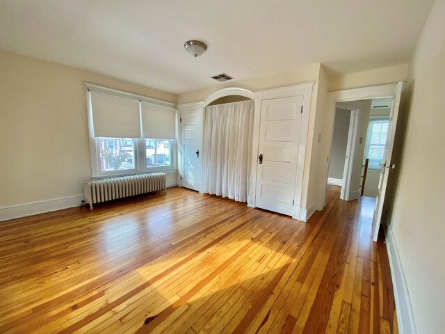 unfurnished bedroom with arched walkways, light wood-style flooring, visible vents, baseboards, and radiator heating unit