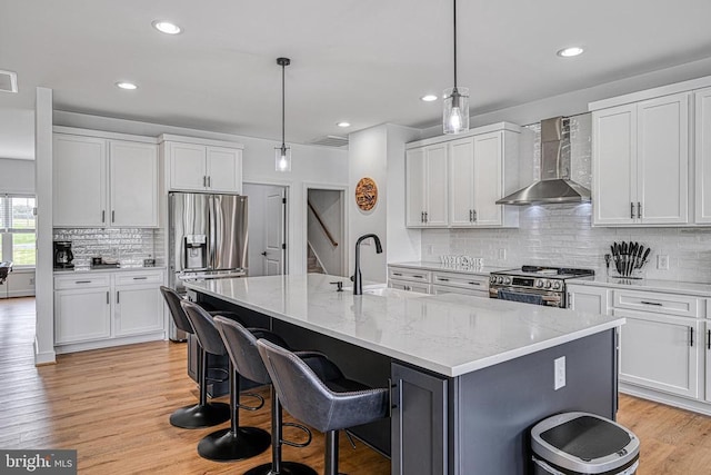 kitchen featuring appliances with stainless steel finishes, an island with sink, white cabinets, hanging light fixtures, and wall chimney range hood