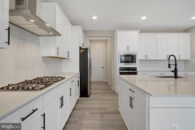 kitchen with light countertops, appliances with stainless steel finishes, white cabinets, a sink, and wall chimney exhaust hood