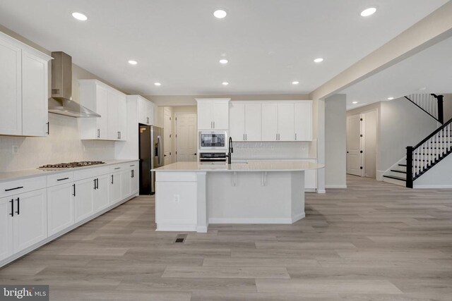 kitchen featuring a center island with sink, stainless steel appliances, light countertops, white cabinetry, and wall chimney range hood