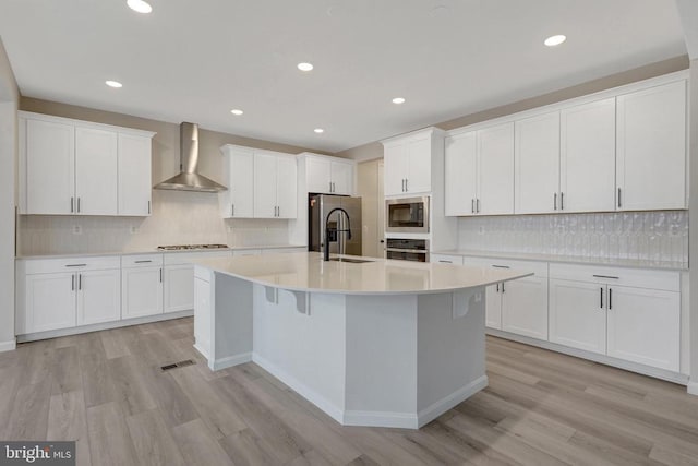 kitchen featuring white cabinets, an island with sink, wall chimney exhaust hood, appliances with stainless steel finishes, and light countertops