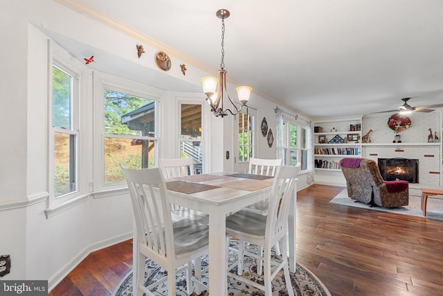 dining space featuring crown molding, dark wood-type flooring, ceiling fan with notable chandelier, and a fireplace