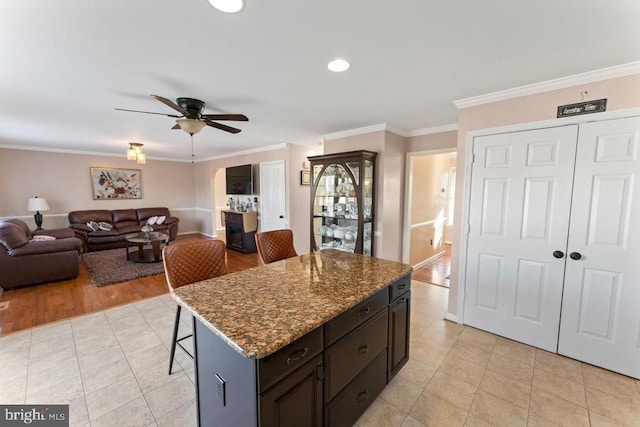 kitchen featuring a kitchen bar, a ceiling fan, dark stone countertops, light tile patterned flooring, and crown molding