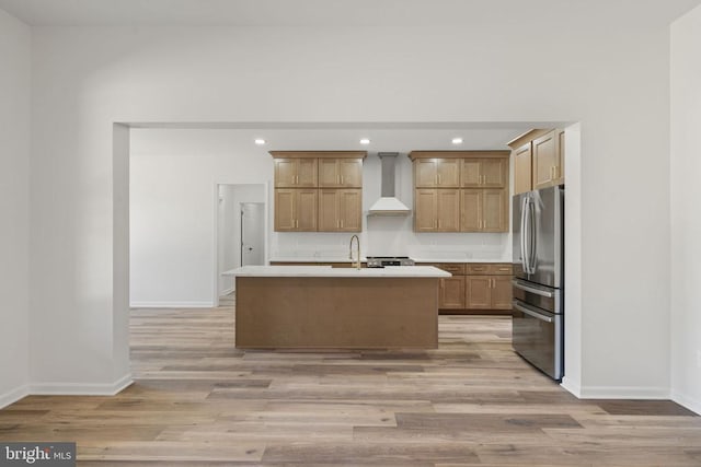 kitchen featuring a kitchen island with sink, wall chimney range hood, light hardwood / wood-style floors, and stainless steel refrigerator