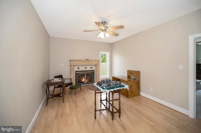 recreation room with a ceiling fan, a warm lit fireplace, light wood-style flooring, and baseboards