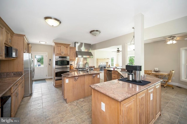 kitchen with a center island with sink, appliances with stainless steel finishes, wall chimney range hood, light stone countertops, and baseboards