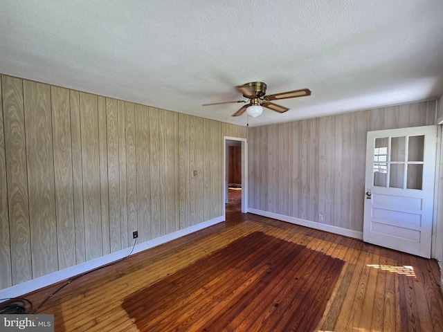 empty room featuring a ceiling fan, a textured ceiling, hardwood / wood-style flooring, baseboards, and wooden walls