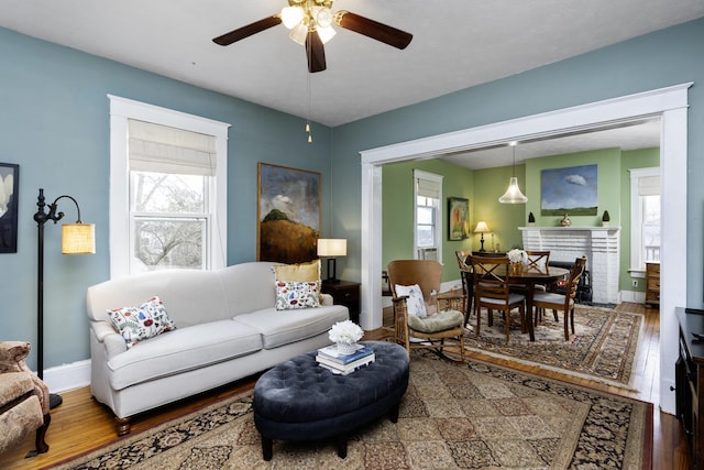 living room with a brick fireplace, a ceiling fan, baseboards, and wood finished floors