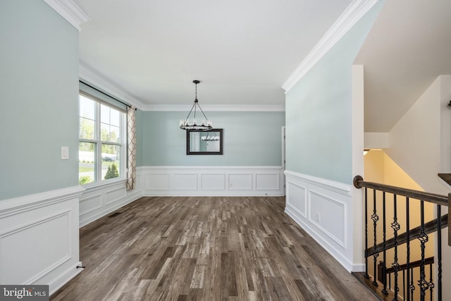 unfurnished dining area featuring crown molding, an inviting chandelier, wainscoting, a decorative wall, and dark wood-type flooring