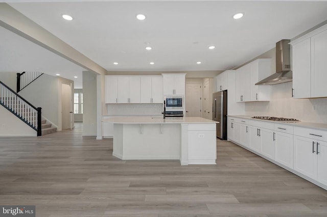 kitchen with stainless steel appliances, white cabinetry, light countertops, wall chimney exhaust hood, and a center island with sink