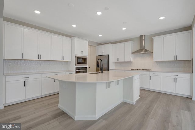 kitchen with an island with sink, wall chimney exhaust hood, stainless steel appliances, light countertops, and white cabinetry