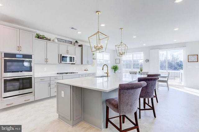 kitchen with a center island with sink, stainless steel appliances, sink, gray cabinets, and pendant lighting