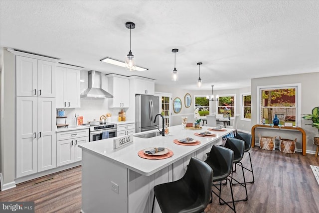 kitchen featuring wall chimney range hood, white cabinetry, a kitchen island with sink, and appliances with stainless steel finishes