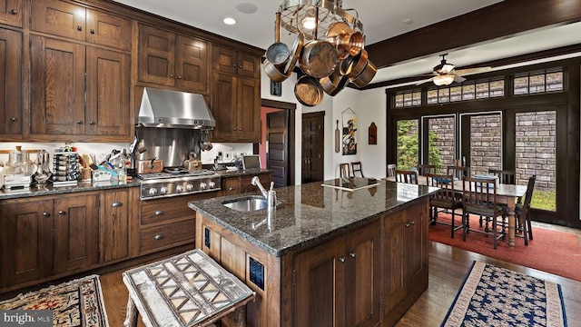 kitchen featuring sink, dark stone countertops, an island with sink, stainless steel gas stovetop, and exhaust hood