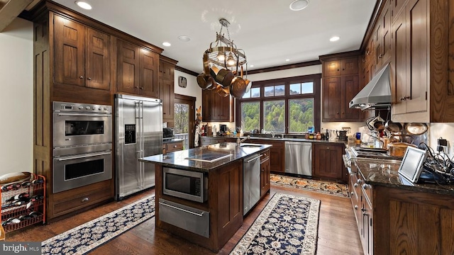 kitchen with a kitchen island, ventilation hood, dark stone counters, built in appliances, and dark brown cabinets