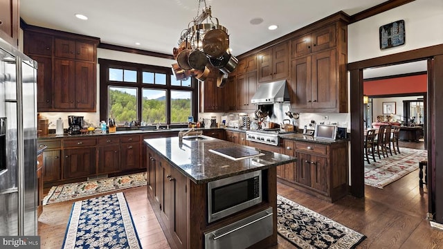 kitchen with wall chimney range hood, sink, appliances with stainless steel finishes, a center island with sink, and dark stone counters