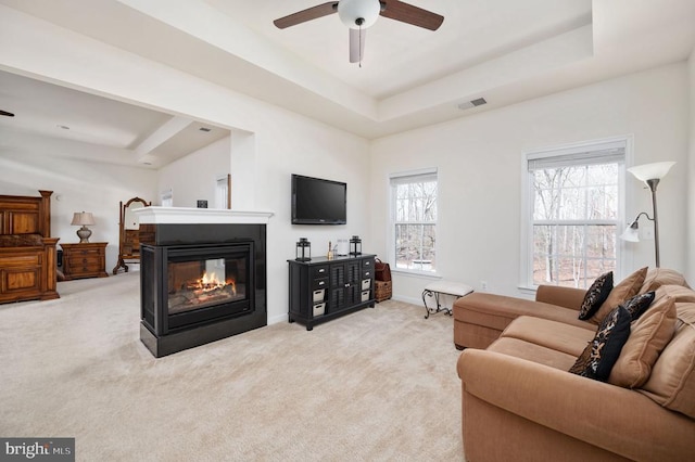 living area featuring visible vents, a multi sided fireplace, a tray ceiling, carpet floors, and a ceiling fan