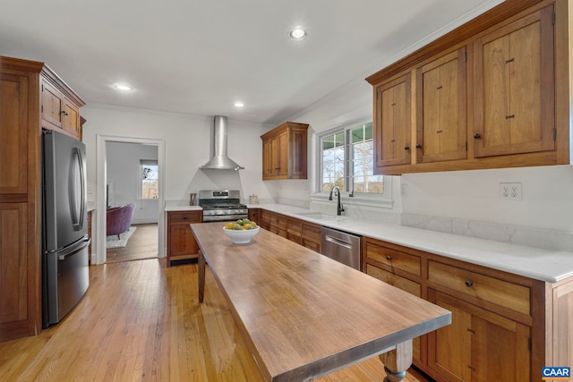 kitchen featuring a sink, light wood-style floors, appliances with stainless steel finishes, brown cabinetry, and wall chimney range hood