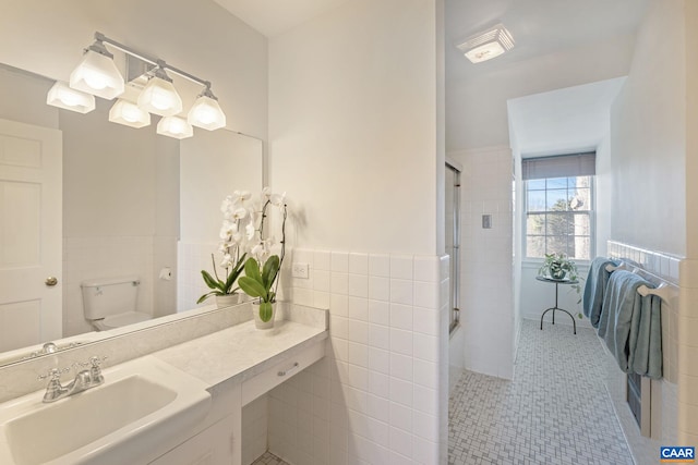 bathroom featuring a shower with shower door, a wainscoted wall, toilet, a sink, and tile walls