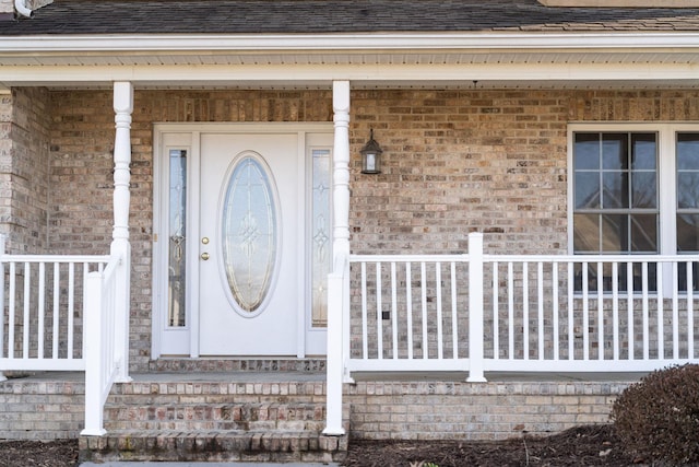 doorway to property with roof with shingles, a porch, and brick siding