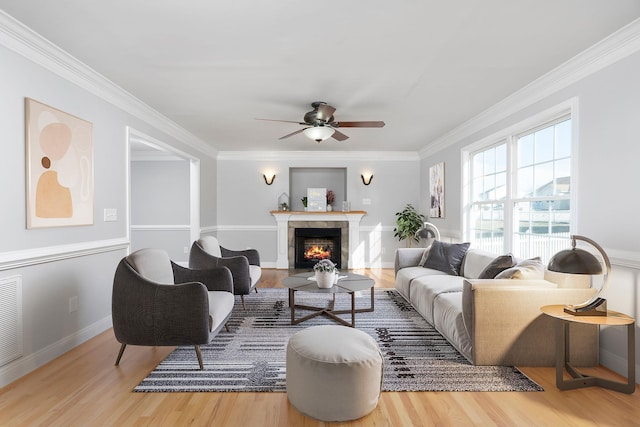 living area featuring a warm lit fireplace, wood finished floors, a ceiling fan, and crown molding