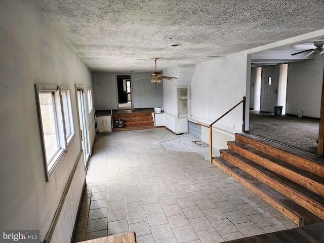 unfurnished living room featuring ceiling fan, a textured ceiling, plenty of natural light, and stairs