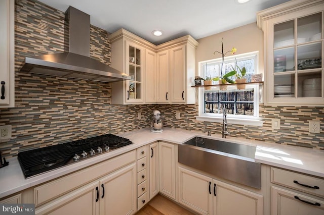 kitchen featuring wall chimney range hood, stainless steel gas cooktop, decorative backsplash, cream cabinetry, and a sink