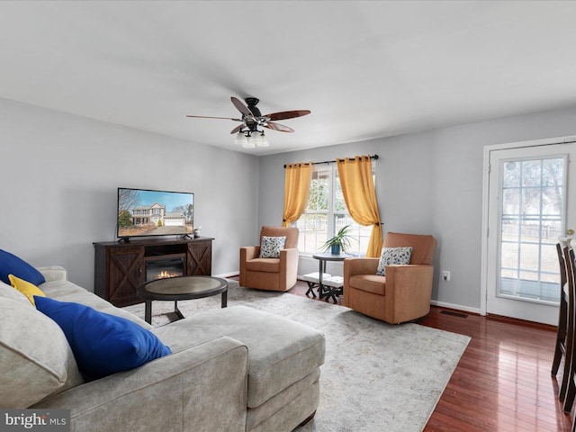 living area with visible vents, dark wood-type flooring, ceiling fan, baseboards, and a glass covered fireplace