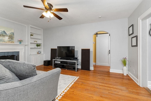 living room with built in features, arched walkways, a fireplace, light wood finished floors, and baseboards