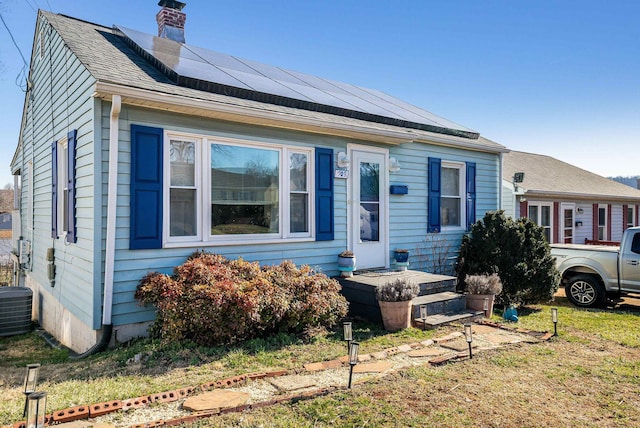 bungalow-style house featuring roof with shingles, central AC unit, roof mounted solar panels, and a chimney