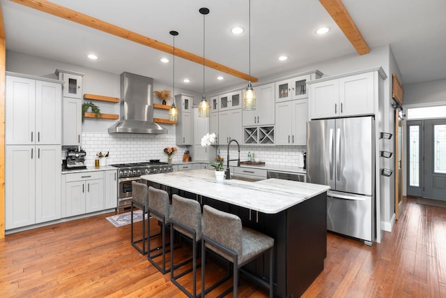 kitchen with light stone counters, appliances with stainless steel finishes, an island with sink, beamed ceiling, and exhaust hood