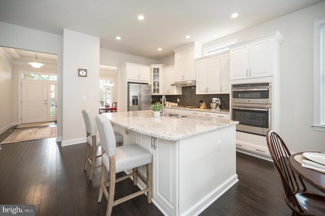 kitchen with appliances with stainless steel finishes, white cabinets, dark hardwood / wood-style flooring, light stone counters, and a center island with sink