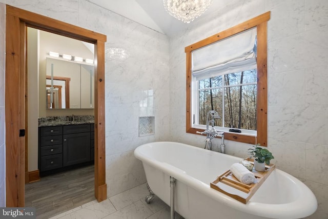 full bath featuring a notable chandelier, tile walls, lofted ceiling, a freestanding bath, and vanity