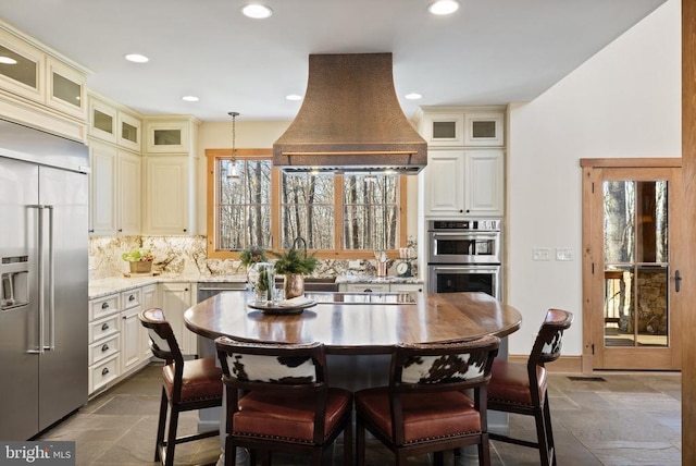 kitchen with stone tile floors, backsplash, appliances with stainless steel finishes, premium range hood, and a kitchen breakfast bar