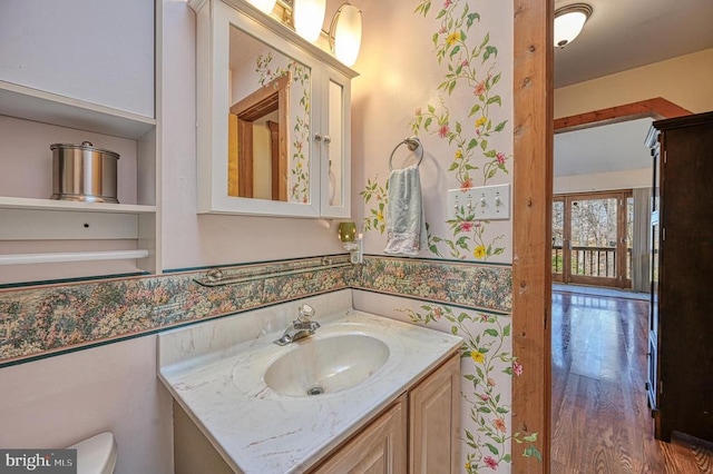 half bathroom featuring wood finished floors, vanity, toilet, and wallpapered walls