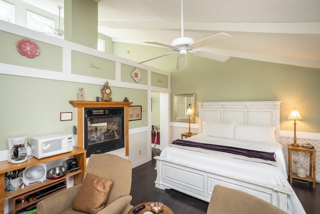 bedroom with vaulted ceiling with beams, wainscoting, a glass covered fireplace, a ceiling fan, and dark wood-style flooring