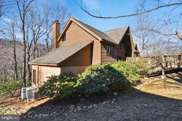 view of side of property featuring a deck, a shingled roof, a chimney, and central AC unit