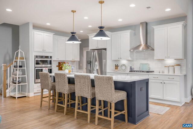 kitchen featuring stainless steel appliances, white cabinets, light countertops, wall chimney range hood, and a center island with sink