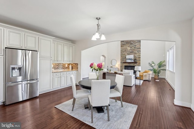 dining area featuring arched walkways, a chandelier, a stone fireplace, baseboards, and dark wood finished floors