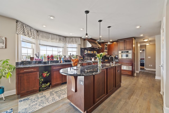 kitchen with oven, a kitchen island with sink, dark stone counters, baseboards, and a warming drawer