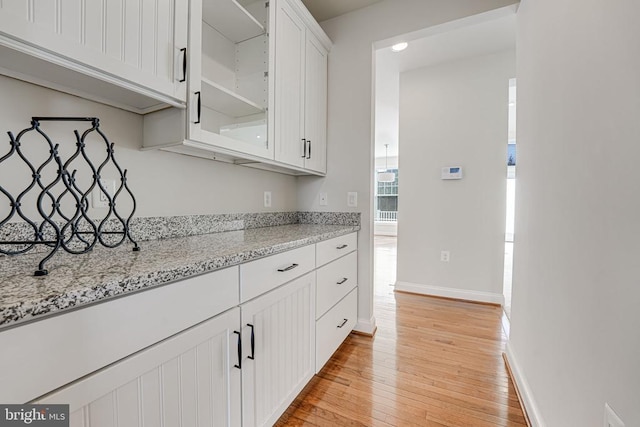 kitchen featuring light wood-type flooring, baseboards, white cabinets, light stone counters, and glass insert cabinets