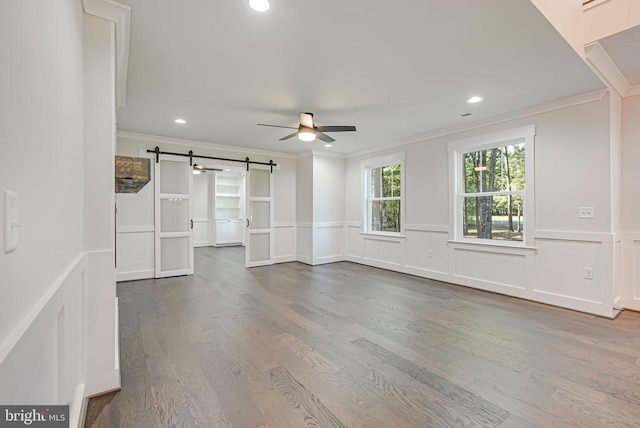 unfurnished living room featuring a ceiling fan, a barn door, ornamental molding, and wood finished floors