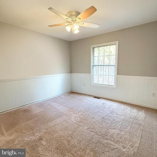 empty room with ceiling fan, visible vents, carpet floors, and a wainscoted wall