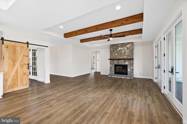 unfurnished living room featuring beam ceiling, a barn door, a stone fireplace, and dark hardwood / wood-style flooring