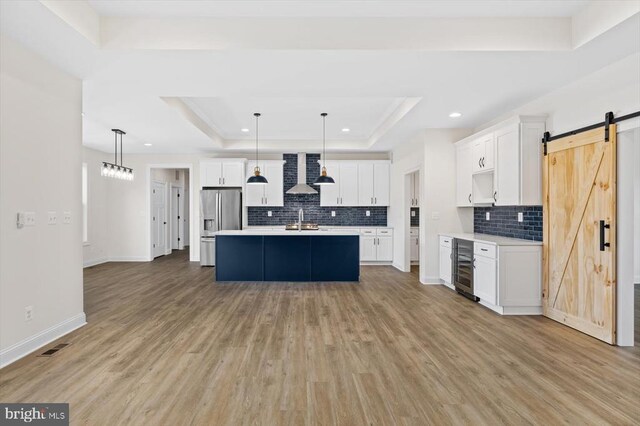 kitchen featuring hanging light fixtures, a tray ceiling, a center island with sink, stainless steel fridge with ice dispenser, and a barn door